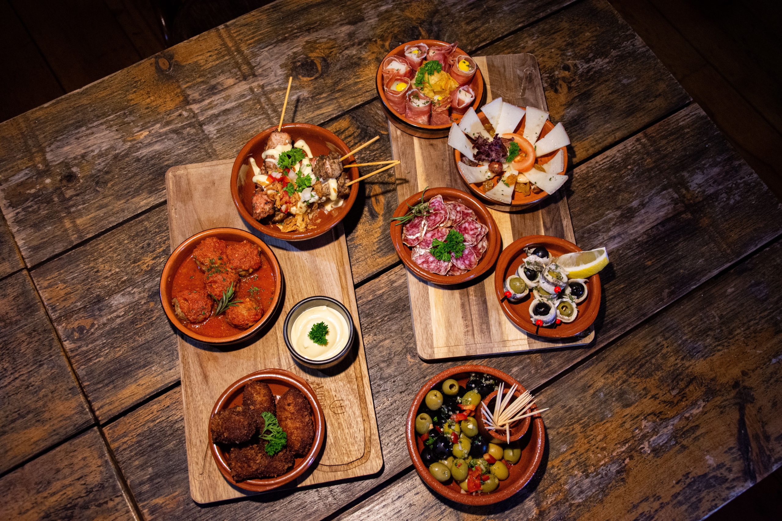 A spread of Spanish tapas — meatballs, croquetas, olives, cured ham and cheese — in terracotta dishes on a dark wooden table
